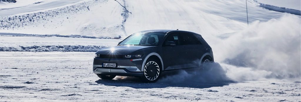 An electric vehicle driving on snow-covered terrain, kicking up snow as it moves.