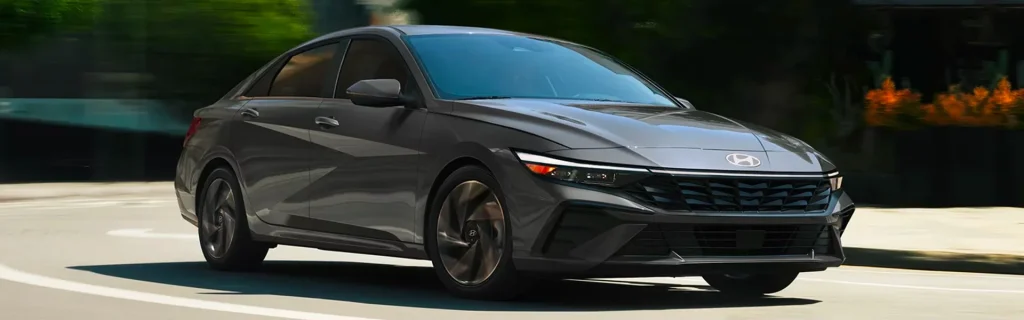 The image shows a sleek, modern 2026 Hyundai Elantra Hybrid driving on a city street. Its aerodynamic design and stylish exterior highlight its performance and efficiency.